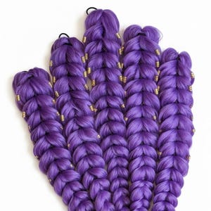 May include: Five vibrant purple braided hair extensions with gold-colored beads. The braids are thick and neatly styled, with a smooth texture. The extensions are arranged against a white background, showcasing the rich color and detail.