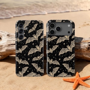 May include: Two black phone cases featuring a repeating pattern of beige bats and stars. The cases are displayed on a sandy beach, with a starfish and driftwood visible in the background.