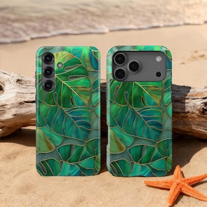 May include: Two phone cases with a vibrant green and gold leaf design. The cases feature a tropical monstera leaf pattern with gold outlines. The cases are on a sandy beach with a starfish and driftwood.