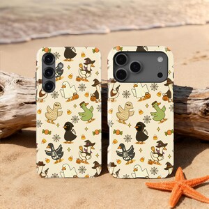 May include: Two phone cases with a Halloween-themed pattern of ducks in costumes, candy corn, and spiderwebs on a cream background. One case has three camera lenses, and the other has four. The cases are on a sandy beach.
