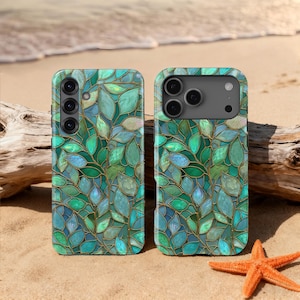 May include: Two phone cases with a stained glass leaf design in shades of green, blue, and gold. The cases are on a sandy beach with a starfish and driftwood in the background.