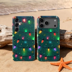May include: Two phone cases with a festive Christmas lights design. The cases are dark green with colorful string lights and small white dots. The cases are on a sandy beach with a starfish and driftwood.