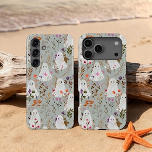 Op de afbeelding: Twee telefoonhoesjes met een herhaald patroon van witte geesten, bloemen en bladeren op een lichtgroene achtergrond. De hoesjes liggen op een zandstrand met een zeester en drijfhout.
