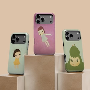 May include: Three phone cases with unique artwork. One case is light green with a girl in a yellow dress, another is pink with a girl in a light blue dress, and the third is light green with a girl with green hair. The cases are on a brown surface.
