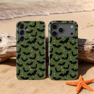 May include: Two phone cases with a green background and a pattern of black bats and white stars. The cases are on a sandy surface with a starfish and driftwood in the background.