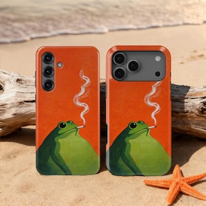 May include: Two phone cases featuring a green frog smoking a cigarette, set against an orange backdrop. The cases are on a sandy beach with a starfish and driftwood. The image has a humorous, cartoon-like style.