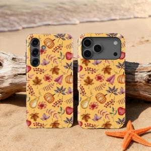 Op de afbeelding: Twee telefoonhoesjes met een gele achtergrond met een herfstthema. Het ontwerp bevat pompoenen, appels, peren, bladeren en bloemen in tinten oranje, rood en paars. De hoesjes liggen op een zandstrand met een zeester.