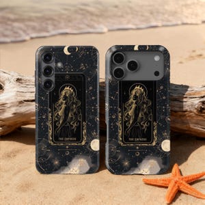 Op de afbeelding: Twee zwarte telefoonhoesjes met een hemels ontwerp met de tarotkaart De Keizerin. De hoesjes hebben een goud- en witkleurenschema met sterren, manen en een portret van een vrouw. Een zeester ligt op het zand.