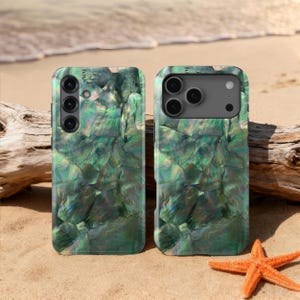 May include: Two phone cases with a shimmering, iridescent green abalone shell design. The cases are displayed on a sandy beach with a starfish and driftwood in the background. The cases have cutouts for the phone's camera.