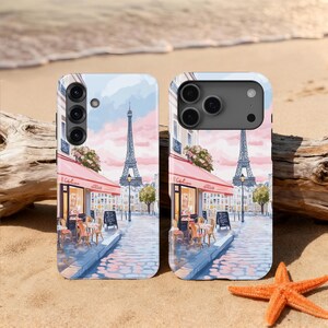 May include: Two phone cases with a watercolor painting of a Parisian cafe scene, featuring the Eiffel Tower, red awnings, and outdoor seating. The cases are on a sandy surface with a starfish and driftwood.