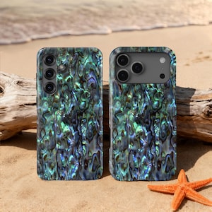 May include: Two phone cases with a shimmering, iridescent abalone shell design in shades of blue, green, and silver. The cases are displayed on a sandy beach with a starfish and driftwood in the background.