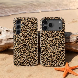 May include: Two mobile phone cases featuring a leopard print pattern in brown and tan. One case has three camera lenses, the other has four. The cases are displayed on a sandy beach with a starfish and driftwood.