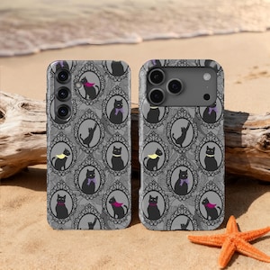 Op de afbeelding: Twee telefoonhoesjes met een herhalend patroon van zwarte katten in sierlijke lijsten op een grijze achtergrond. De katten dragen kleurrijke accessoires. De hoesjes liggen op een zandstrand met een zeester en drijfhout.