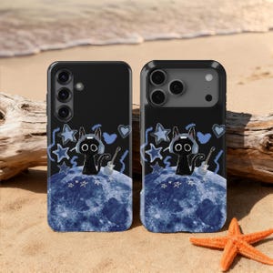 May include: Two black phone cases with a whimsical design. Each case features a black cat wearing headphones and holding a guitar, set against a blue moon and surrounded by stars and hearts. The cases are on a sandy beach.