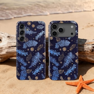 May include: Two phone cases with a navy blue background and a pattern of blue and gold pine needles, pine cones, and floral accents. The cases are on a sandy beach with a starfish and driftwood.