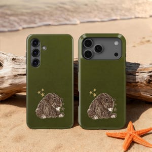 Può includere: Due custodie per telefono verde oliva con un'illustrazione di orso e dettagli a stella. L'orso è marrone e bianco. Le custodie sono su una spiaggia sabbiosa con una stella marina e legno alla deriva.