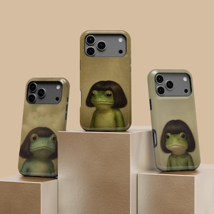 May include: Three phone cases with a whimsical illustration of a frog wearing a dark brown wig. The cases have a tan background and are displayed on a tiered, neutral-toned platform. The frog has large eyes and a green body.