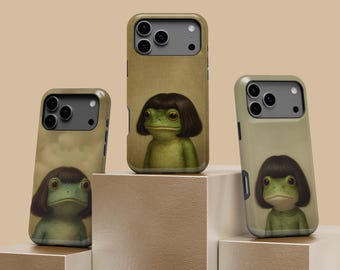 May include: Three phone cases featuring a quirky illustration of a frog with a dark brown wig. The cases have a tan background and are displayed on a tiered, neutral-toned platform. The frog has large eyes and a green body.
