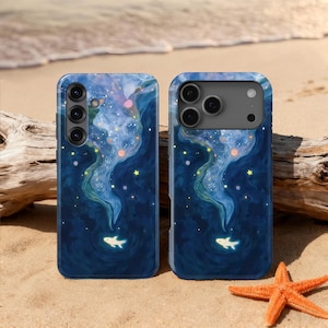 Puede incluir: Dos fundas para teléfono con un diseño azul oscuro con temática de galaxia. Las fundas presentan una nebulosa arremolinada con estrellas y un pez brillante en la parte inferior. Las fundas están en una playa de arena con una estrella de mar y madera flotante.