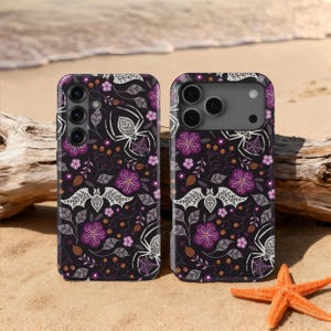 Könnte beinhalten: Zwei schwarze Handyhüllen mit einem sich wiederholenden Muster aus weißen Spinnen, Fledermäusen und lila Blumen. Die Hüllen befinden sich an einem Sandstrand mit einem Seestern und Treibholz im Hintergrund. Die Hüllen haben unterschiedliche Kameradesigns.
