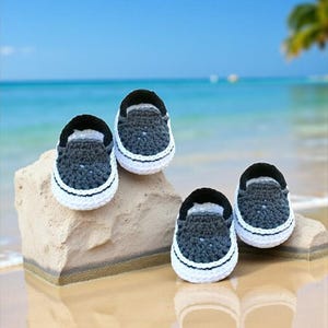 May include: Four pairs of baby crochet shoes in a slip-on style. The shoes are dark gray with black trim and white soles. They are displayed on a light-colored rock with a beach and ocean background.