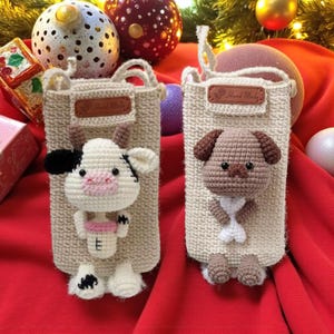 May include: Two beige crochet phone cases, one with a cow and the other with a dog. The cow has black and white spots, a pink nose, and holds a pink cup. The dog has brown ears and a white bow. Each case has a "Hand Made" label.