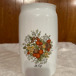 May include: White ceramic canister with a floral design featuring coffee beans, nuts, and berries in shades of brown, orange, and yellow. The words "Le Café" are printed below the design. The canister has a cylindrical shape.