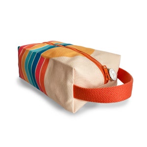Op de afbeelding: Een beige canvas reistas met een ritssluiting en een oranje handvat. De tas heeft een kleurrijk gestreept patroon in tinten blauw, rood, oranje en geel.