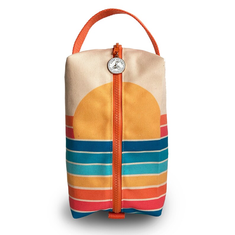 Op de afbeelding: Een beige canvas tas met rits met een zonsondergangontwerp. De tas heeft een bovenste handvat en een ritssluiting. De rits is oranje en het ontwerp heeft een gele zon, blauwe en turquoise strepen en oranje en roze strepen.