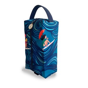 Op de afbeelding: Een blauwe toilettas met rits, een golfpatroon en een afbeelding van een vrouw die surft. De tas heeft een bovenste handvat.