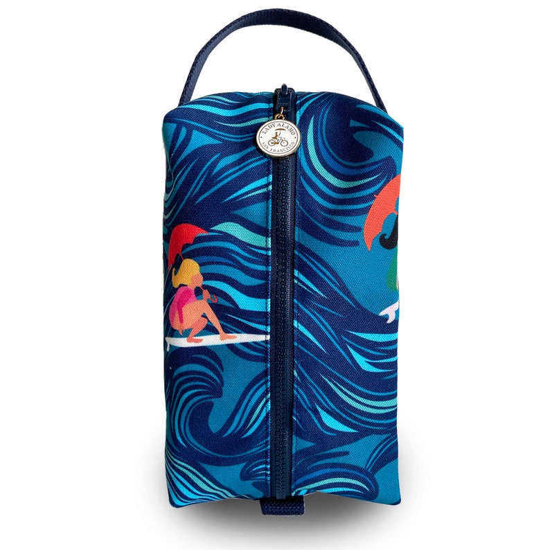 Op de afbeelding: Een blauwe ritszak met een golfpatroon en een illustratie van een surfermeisje. De tas heeft een donkerblauwe handgreep en een gouden rits met een logo dat "Lady Alamo, San Francisco" zegt.