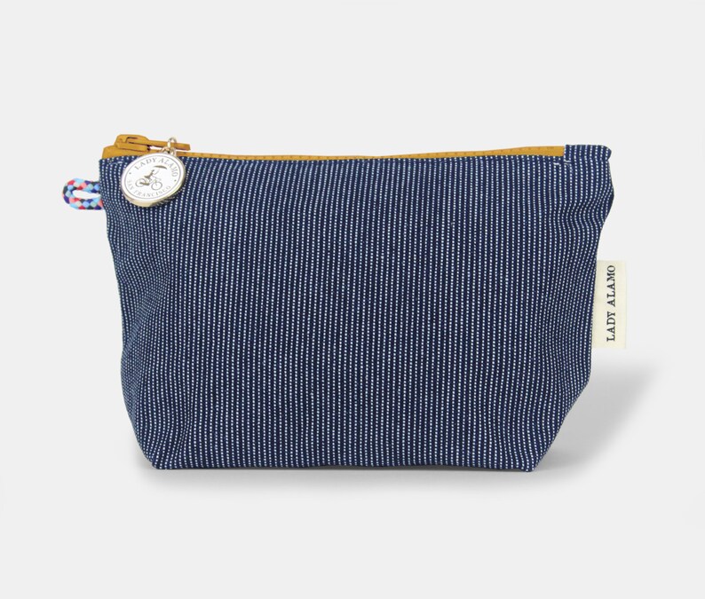 Puede incluir: Una peque&ntilde;a bolsa de cremallera a rayas azules y blancas con un tirador de cremallera de cuero marr&oacute;n y un charm plateado. El charm tiene el texto "Lady Alamo" en &eacute;l. La bolsa est&aacute; hecha de una tela suave y duradera y es perfecta para llevar objetos peque&ntilde;os.