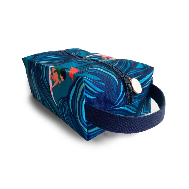 Op de afbeelding: Een blauwe toilettas met een ritssluiting en een donkerblauwe handgreep. De tas heeft een kleurrijk printje van surfers die op golven rijden.