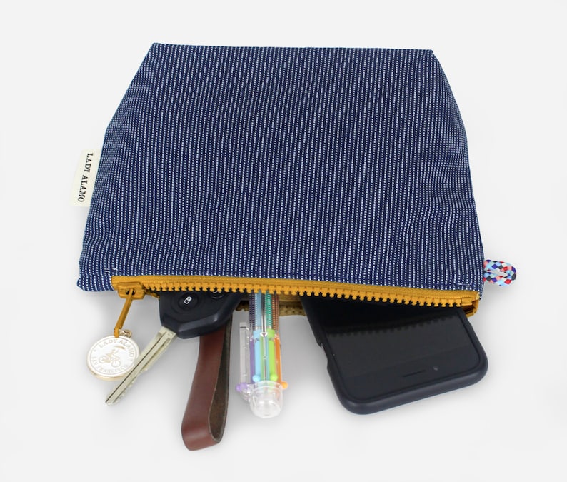 Puede incluir: Una bolsa de cremallera a rayas azules y blancas con una cremallera amarilla. La bolsa est&aacute; abierta y contiene un tel&eacute;fono, llaves, un bol&iacute;grafo y un llavero. La bolsa tiene el texto "LADY ALAMO" impreso en el lateral.