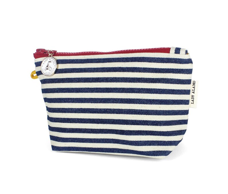 Puede incluir: Una bolsa de maquillaje de lona a rayas azules y blancas con una cremallera roja y un dije plateado con un dise&ntilde;o de bicicleta. La bolsa tiene el texto "LADY ALAMO" impreso en el lateral.