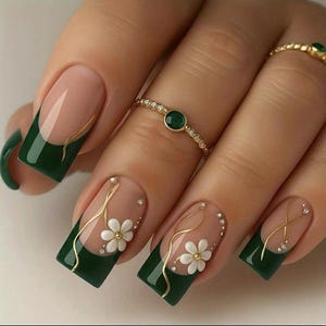 May include: Manicured nails with a dark green French tip design, adorned with gold accents, white flower embellishments, and small rhinestones. A gold ring with a green gemstone is also visible.