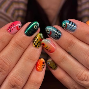 Puede incluir: Un conjunto de u&ntilde;as con dise&ntilde;os detallados de arte de u&ntilde;as que representan varios peces. Cada u&ntilde;a muestra una paleta de colores &uacute;nica que incluye rosa, verde met&aacute;lico, amarillo con puntos negros, azul, naranja y verde oliva, con ojos pintados.