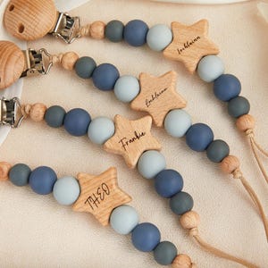 May include: Wooden baby pacifier clips with star-shaped name tags. The clips feature a combination of wooden, blue, and gray beads. Each star is engraved with a name: Theo, Frankie, and Lochlainn. The pacifier clips have a wooden clip and a cord for attaching to a pacifier.