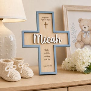 May include: A wooden cross with a light blue frame and a natural wood centre. The cross features the name Micah in white 3D lettering, the text Baptised April 6, 2026, a small cross icon, and the quote Walk in faith and love each day. Psalm 119:105.