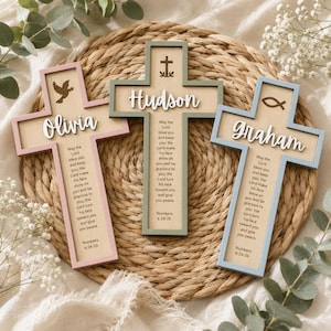 Könnte beinhalten: Drei Holzkreuze mit hellen Holzkernen und farbigen Rändern in Rosa, Salbeigrün und Hellblau. Jedes Kreuz zeigt einen Namen in weißer Schreibschrift, ein religiöses Symbol und den Text aus Numeri 6,24-26 in schwarzer Schrift auf dem Holz.