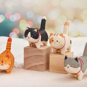 May include: Four wooden cat figurines in various colors: orange, black and white, cream and brown, and gray and white. Each cat has a bell collar and is standing on a small wooden block. The cats have smiling faces and are hand-carved.