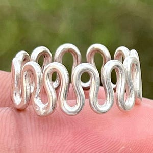 Może przedstawiać: Biały ceramiczny kubek z czarnym uchwytem i napisem "But First, Coffee" pogrubioną, czarną czcionką. Kubek jest ustawiony na rozmytym tle, sugerującym przytulne otoczenie.