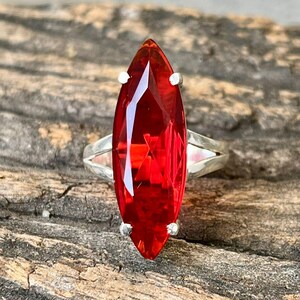 Peut inclure: Une bague en argent ornée d'une grande pierre rouge vif taillée en marquise. La pierre est sertie de quatre griffes et présente une surface à facettes. La bague est présentée sur une surface en bois vieilli.