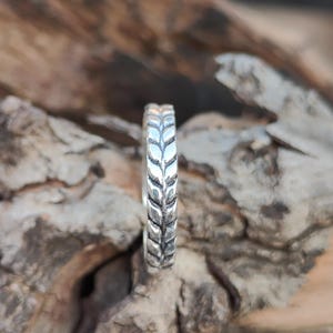 May include: A silver ring featuring a continuous embossed wheat or leaf pattern around the entire band. The metal has a polished finish with darkened recessed areas that highlight the intricate, repeating botanical design along the outer surface.