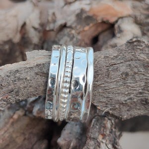 Puede incluir: Un anillo de color plateado con un diseño de varias bandas, que presenta una banda central texturizada flanqueada por dos bandas más anchas con acabado martillado. La superficie metálica es pulida y reflectante, con hendiduras marcadas.