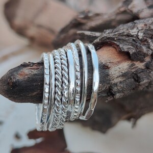 May include: A set of seven silver-toned stacking rings featuring various textures, including smooth, twisted rope, and hammered finishes. The rings are arranged side-by-side, showcasing their distinct metallic patterns and reflective surfaces.
