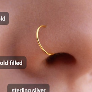Puede incluir: Primer plano de una nariz con un aro de oro. La imagen incluye texto que dice "22k gold", "22k gold filled" y "sterling silver". El aro de la nariz tiene un diseño simple y curvado.