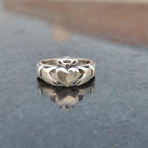 May include: A silver Claddagh ring, a traditional Irish design. It features a heart held by two hands, with a crown above. The ring has intricate detailing and a polished finish. The Claddagh ring symbolises love, loyalty, and friendship.