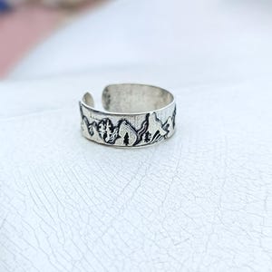 Peut inclure: Bague réglable argentée avec un motif de montagnes et d'arbres. La bague présente un motif de paysage détaillé autour de l'anneau, avec une finition texturée. La bague est ouverte en bas.