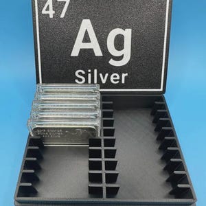 May include: A black storage box with a lid featuring the periodic table symbol for silver, "Ag". The box is open, revealing slots for storing silver bars. Several clear silver bars are visible inside the box.